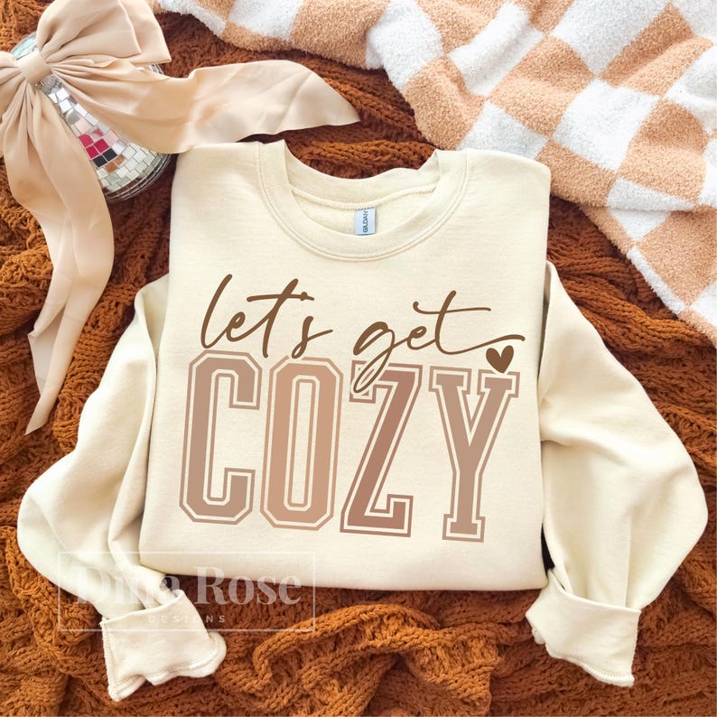 May include: Cream-colored sweatshirt with the phrase "let's get COZY" in brown lettering. The word "COZY" is in a block letter style. The sweatshirt is displayed on a brown knitted blanket with a peach-colored bow and a checkered blanket in the background.