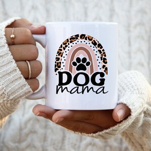 May include: White ceramic mug with a rainbow design featuring a paw print in the center. The rainbow is decorated with a leopard print pattern. The text "DOG mama" is printed below the rainbow.