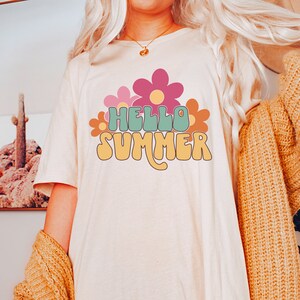 May include: A white t-shirt with a retro-style graphic that says "Hello Summer" in green, yellow, and orange letters. The graphic is surrounded by pink, orange, and yellow flowers.