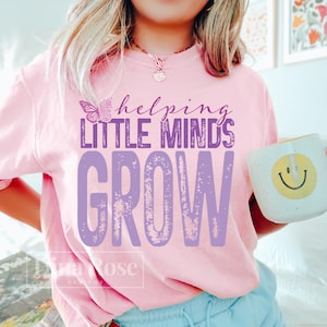 Pu&ograve; includere: Maglietta rosa chiaro con la scritta viola "helping LITTLE MINDS GROW". Una grafica a farfalla &egrave; sopra le parole "LITTLE MINDS". Una tazza bianca con una faccina gialla sorridente &egrave; tenuta in mano.