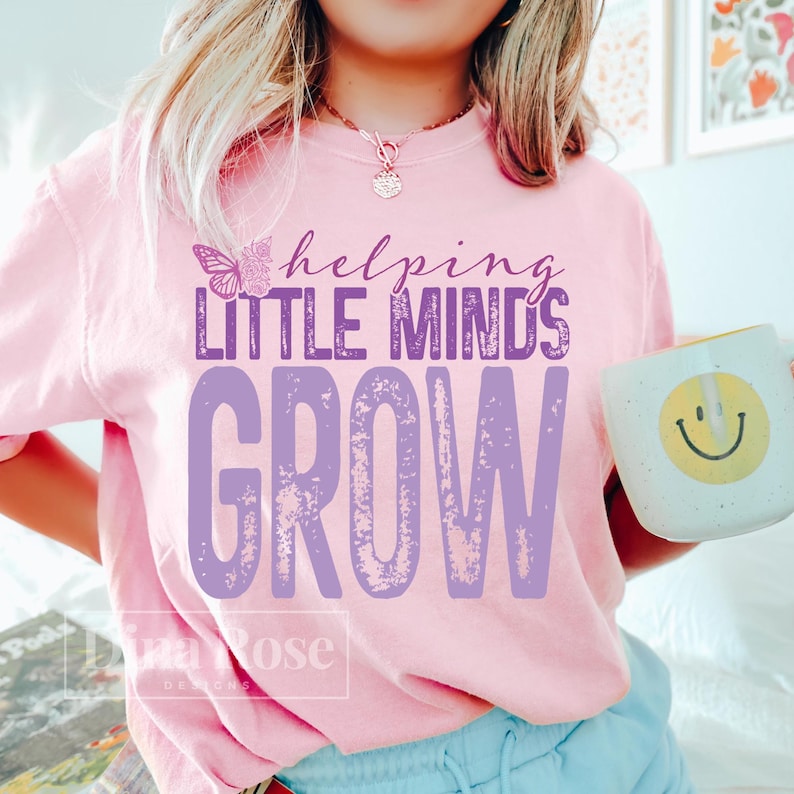 Pu&ograve; includere: Maglietta rosa chiaro con la scritta viola "helping LITTLE MINDS GROW". Una grafica a farfalla &egrave; sopra le parole "LITTLE MINDS". Una tazza bianca con una faccina gialla sorridente &egrave; tenuta in mano.