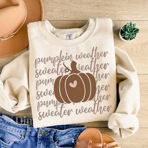 May include: Cream-colored sweatshirt with the text "Pumpkin Weather Sweater Weather" and a brown pumpkin graphic with a heart. The sweatshirt is folded, with a pair of blue jeans and sandals visible.