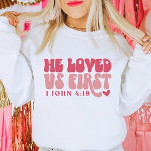 May include: White sweatshirt with a pink and red graphic that says "He Loved Us First" and "1 John 4:19".