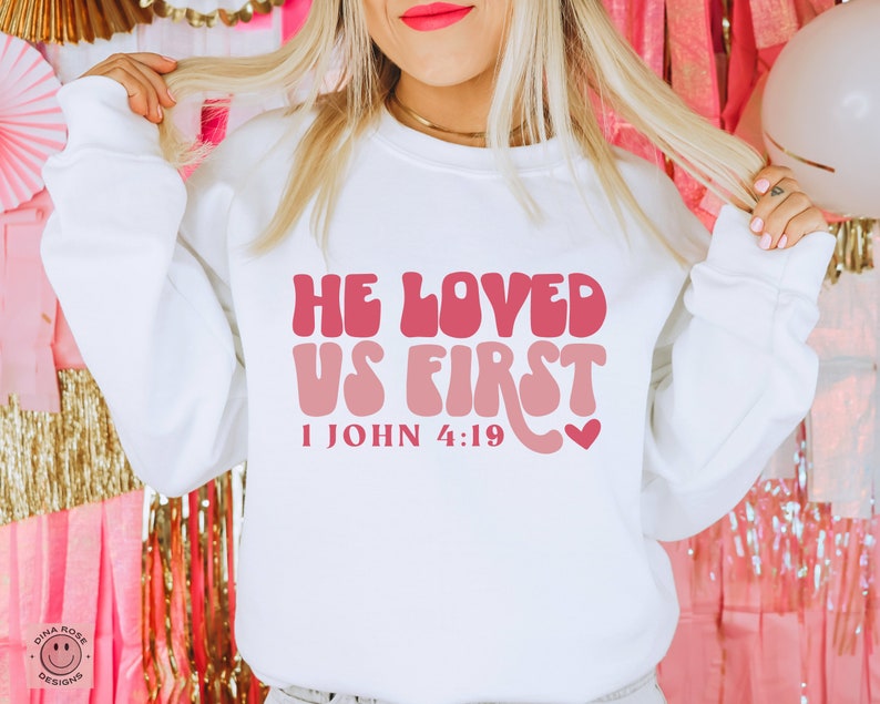 May include: White sweatshirt with a pink and red graphic that says "He Loved Us First" and "1 John 4:19".