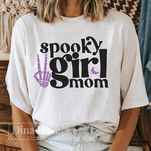 May include: White t-shirt with the text "spooky girl mom" in black. A purple skeleton hand with a peace sign is to the left of the text.