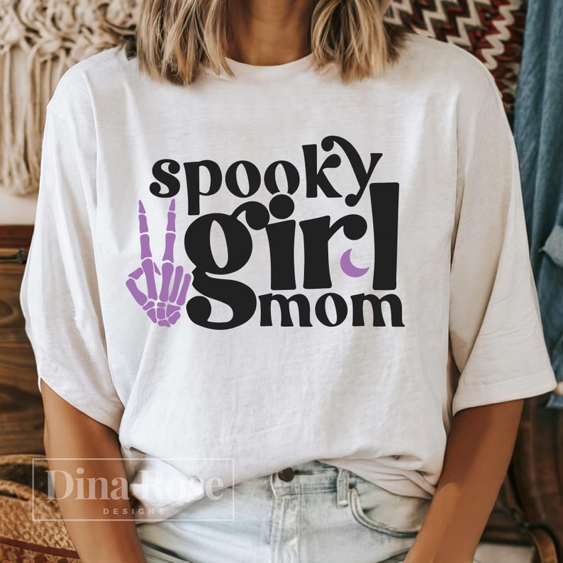 May include: White t-shirt with the text "spooky girl mom" in black. A purple skeleton hand with a peace sign is to the left of the text.