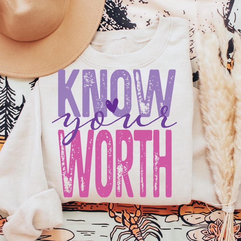 K&ouml;nnte beinhalten: Ein wei&szlig;es Sweatshirt mit den Worten "KNOW your WORTH" in lila und rosa Buchstaben. Das Sweatshirt ist gefaltet, wobei die &Auml;rmel sichtbar sind. Ein beigefarbener Hut und dekorative Elemente befinden sich im Hintergrund.