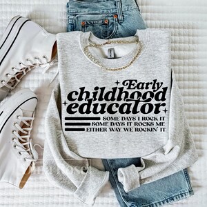 以下が含まれることがあります： 「Early childhood educator」の文字が黒でプリントされたライトグレーのスウェットシャツ。スウェットシャツには、「Some days I rock it」、「Some days it rocks me」、「Either way we rockin' it」というフレーズも含まれています。スウェットシャツは、青いジーンズと白いスニーカーと組み合わせています。