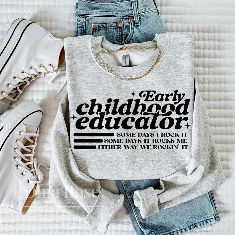 以下が含まれることがあります： 「Early childhood educator」の文字が黒でプリントされたライトグレーのスウェットシャツ。スウェットシャツには、「Some days I rock it」、「Some days it rocks me」、「Either way we rockin' it」というフレーズも含まれています。スウェットシャツは、青いジーンズと白いスニーカーと組み合わせています。
