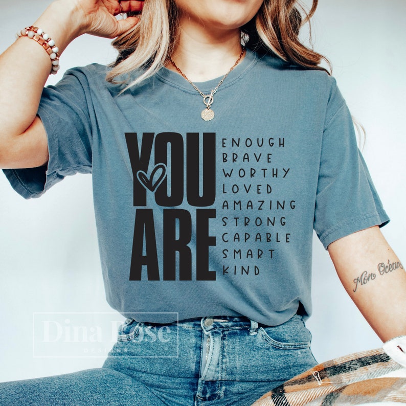 May include: A blue t-shirt with the words "YOU ARE" in large black letters. The words "ENOUGH, BRAVE, WORTHY, LOVED, AMAZING, STRONG, CAPABLE, SMART, KIND" are arranged around the words "YOU ARE" in a word search style.