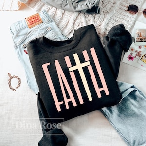 May include: A black sweatshirt with the word "FAITH" in a light pink, block letter font, and a light yellow cross. The sweatshirt is displayed with a pair of blue jeans, a bracelet, and sunglasses.