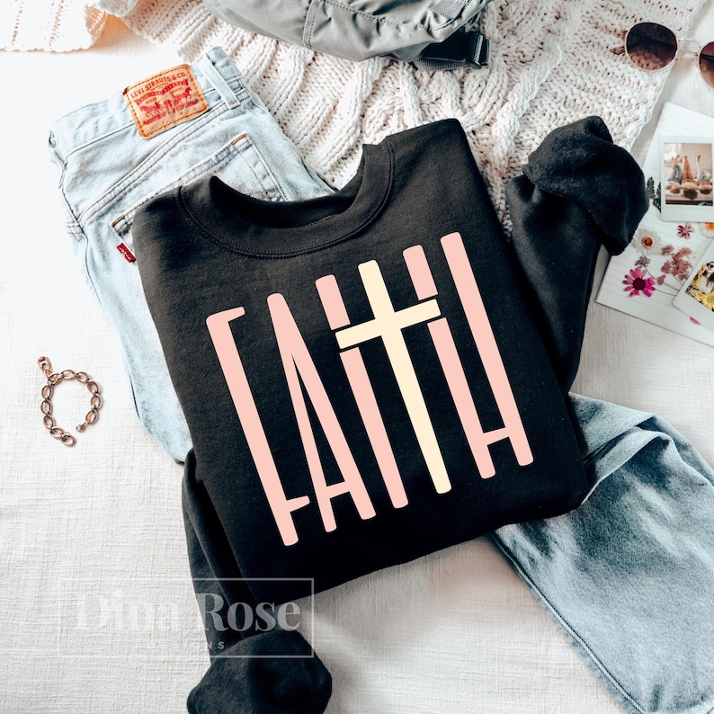 May include: A black sweatshirt with the word "FAITH" in a light pink, block letter font, and a light yellow cross. The sweatshirt is displayed with a pair of blue jeans, a bracelet, and sunglasses.