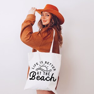 May include: A white tote bag with the text "Life is better at the beach" in black lettering. The bag features a graphic of a sun setting over waves. A woman is holding the bag and wearing a brown sweater and a brown hat.