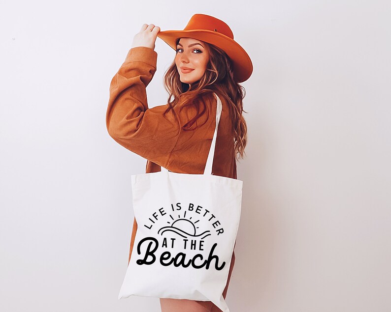 May include: A white tote bag with the text "Life is better at the beach" in black lettering. The bag features a graphic of a sun setting over waves. A woman is holding the bag and wearing a brown sweater and a brown hat.