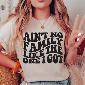 Pu&ograve; includere: Una maglietta bianca con una grafica nera che recita "Ain't no family like the one I got" in un font retr&ograve;.