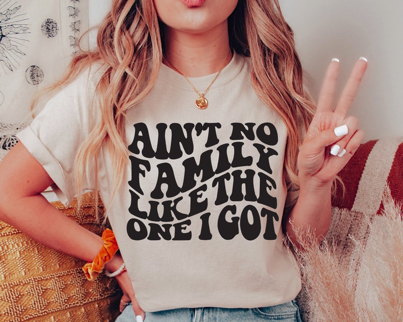 Pu&ograve; includere: Una maglietta bianca con una grafica nera che recita "Ain't no family like the one I got" in un font retr&ograve;.