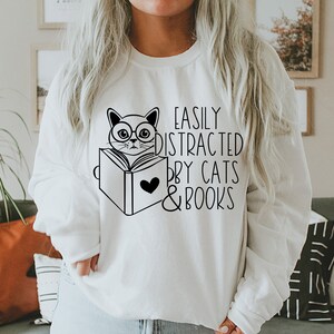 K&ouml;nnte beinhalten: Wei&szlig;er Pullover mit einer schwarz-wei&szlig;en Illustration einer Katze mit Brille, die ein Buch liest. Der Text "Easily Distracted by Cats & Books" ist auf dem Pullover aufgedruckt.