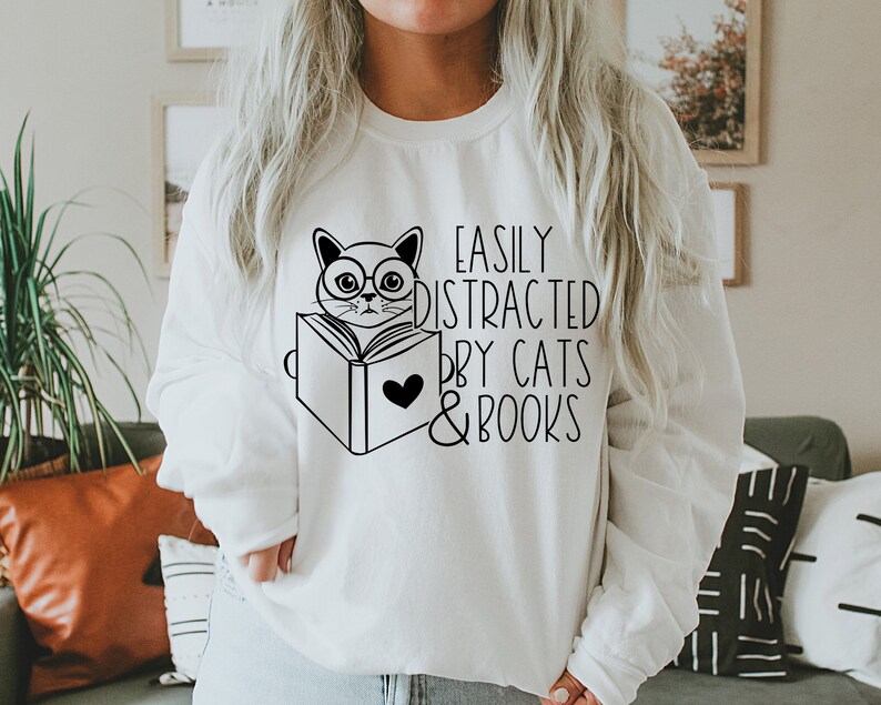 K&ouml;nnte beinhalten: Wei&szlig;er Pullover mit einer schwarz-wei&szlig;en Illustration einer Katze mit Brille, die ein Buch liest. Der Text "Easily Distracted by Cats & Books" ist auf dem Pullover aufgedruckt.