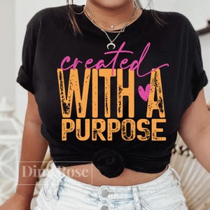 K&ouml;nnte beinhalten: Schwarzes T-Shirt mit den Worten "created WITH A PURPOSE" in orangefarbenen und rosa Buchstaben. Ein rosa Herz befindet sich neben dem Wort "A". Das T-Shirt wird von einer Person getragen.
