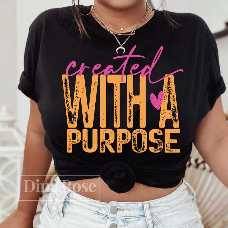 K&ouml;nnte beinhalten: Schwarzes T-Shirt mit den Worten "created WITH A PURPOSE" in orangefarbenen und rosa Buchstaben. Ein rosa Herz befindet sich neben dem Wort "A". Das T-Shirt wird von einer Person getragen.