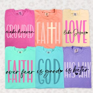 May include: Six colorful t-shirts with inspirational phrases. The shirts are in shades of pink, peach, yellow, teal, blue, and purple. Each shirt features a different text design in contrasting colors, including phrases like "Crowded make heaven" and "Love like Jesus."