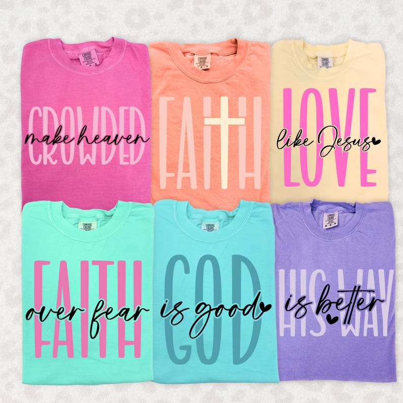 May include: Six colorful t-shirts with inspirational phrases. The shirts are in shades of pink, peach, yellow, teal, blue, and purple. Each shirt features a different text design in contrasting colors, including phrases like "Crowded make heaven" and "Love like Jesus."