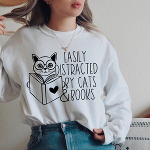 K&ouml;nnte beinhalten: Wei&szlig;er Pullover mit einer schwarzen Illustration einer Katze mit Brille, die ein Buch liest. Der Text "Easily Distracted by Cats & Books" ist auf dem Pullover aufgedruckt.