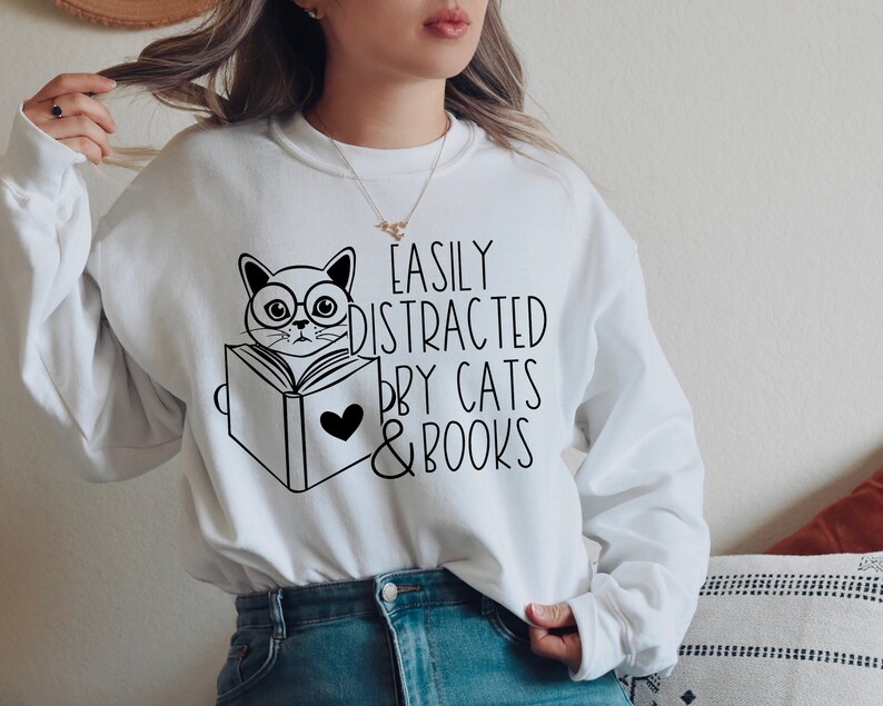 K&ouml;nnte beinhalten: Wei&szlig;er Pullover mit einer schwarzen Illustration einer Katze mit Brille, die ein Buch liest. Der Text "Easily Distracted by Cats & Books" ist auf dem Pullover aufgedruckt.