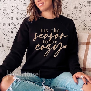 Puede incluir: Sudadera negra con la frase "tis the season to be cozy" en fuente cursiva. La sudadera es de cuello redondo. La imagen tambi&eacute;n muestra a una persona que lleva la sudadera.