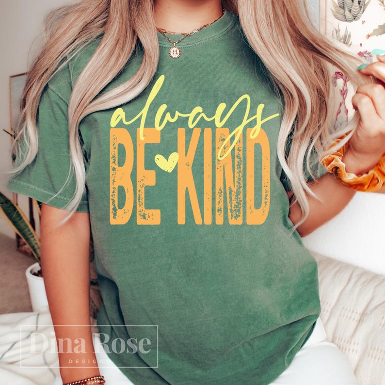 May include: A green t-shirt with the words "always BE KIND" in yellow script and block letters. A yellow heart is between the words "BE" and "KIND". The shirt is a casual style.