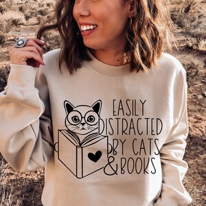 K&ouml;nnte beinhalten: Ein beiger Pullover mit einem schwarzen Motiv eines Katzen mit Brille, das ein Buch liest. Der Text "Easily Distracted by Cats & Books" ist unter der Katze aufgedruckt.