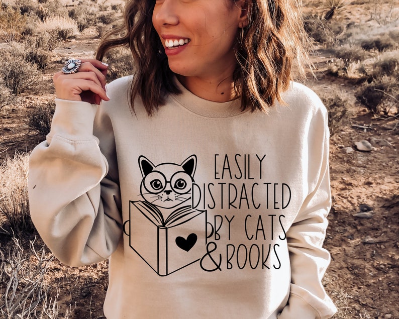 K&ouml;nnte beinhalten: Ein beiger Pullover mit einem schwarzen Motiv eines Katzen mit Brille, das ein Buch liest. Der Text "Easily Distracted by Cats & Books" ist unter der Katze aufgedruckt.
