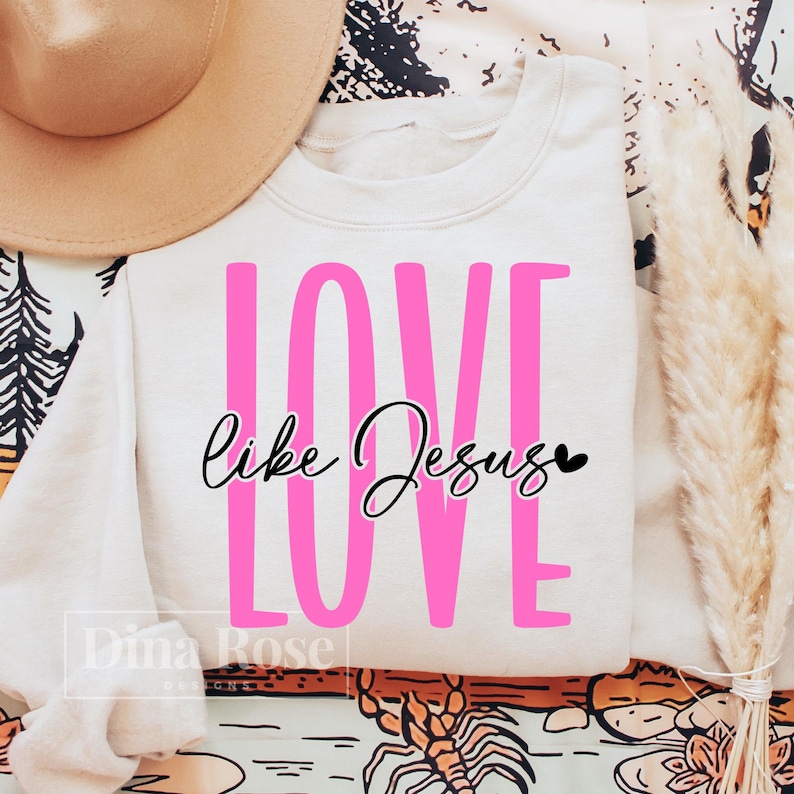 May include: A cream-colored sweatshirt with the words "LOVE" in large pink letters and "like Jesus" in cursive script. A small black heart is next to the word "Jesus". A tan hat is visible in the upper left corner.