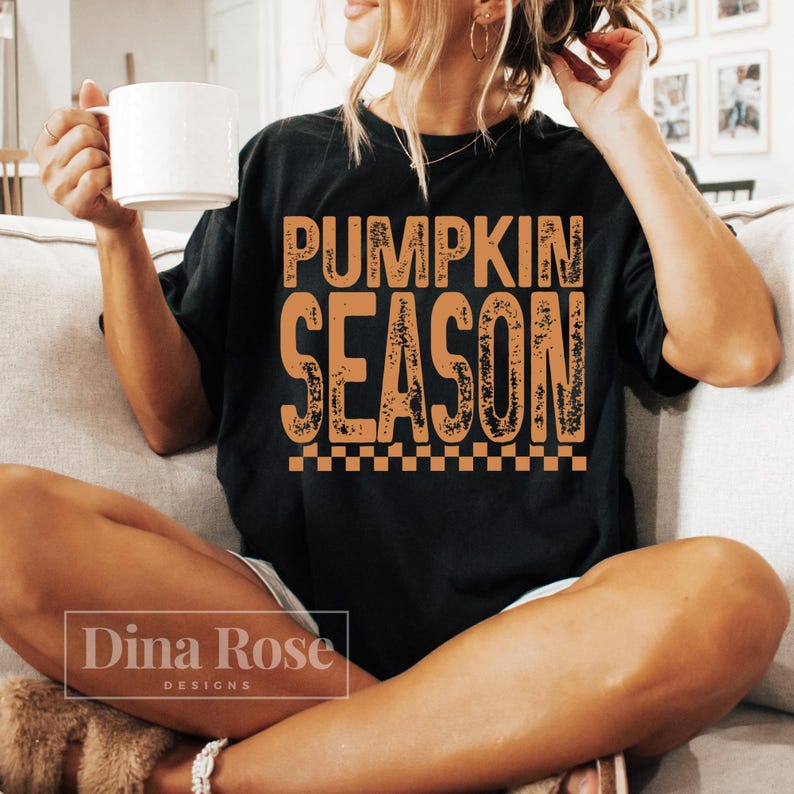 May include: Black t-shirt with the words "PUMPKIN SEASON" in orange, distressed lettering. The shirt has a checkered border at the bottom. A white mug is held in the left hand. The shirt is part of the Dina Rose Designs collection.