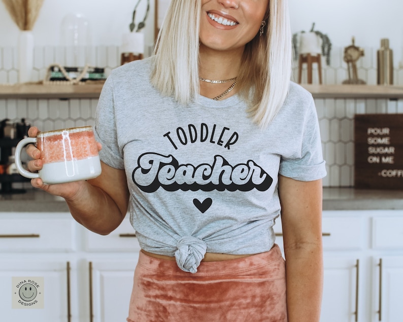 Pu&ograve; includere: Una maglietta grigia con il testo "Toddler Teacher" in un font retr&ograve;. Il testo &egrave; nero e la maglietta ha un piccolo cuore nero sotto il testo.