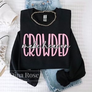 May include: A black sweatshirt with the words "CROWDED make heaven" in pink and white lettering. A gold chain necklace is draped on the sweatshirt, which is placed on top of blue jeans. White sneakers are visible in the background.