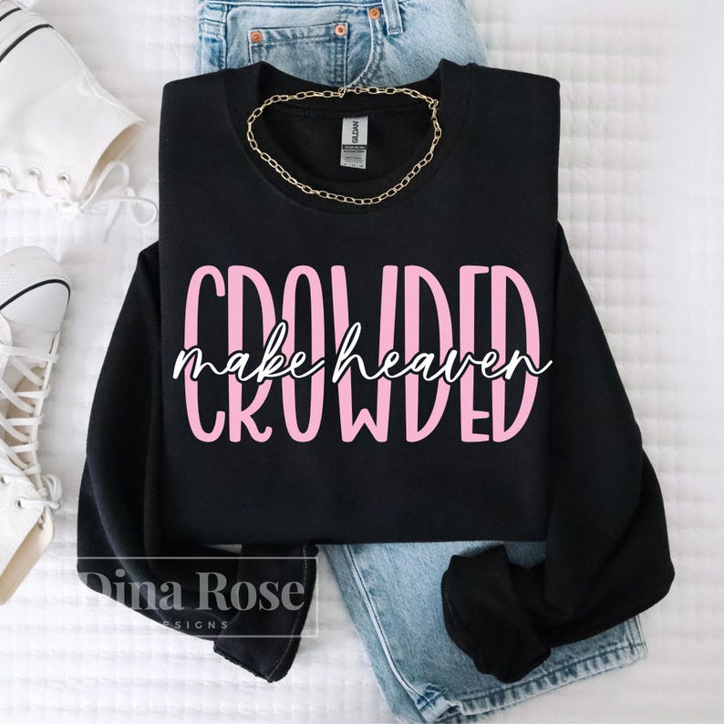 May include: A black sweatshirt with the words "CROWDED make heaven" in pink and white lettering. A gold chain necklace is draped on the sweatshirt, which is placed on top of blue jeans. White sneakers are visible in the background.