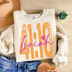 May include: Cream-colored sweatshirt with the text "41:10 Isaiah" in orange and pink. The design features a heart symbol. The sweatshirt is a casual style, suitable for everyday wear.