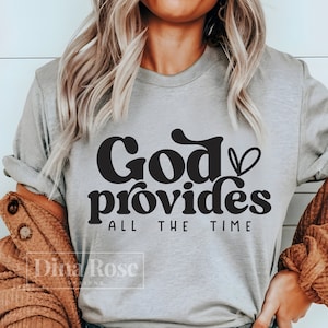 Puede incluir: Una camiseta gris con el texto "God provides all the time" en negro con un símbolo de corazón después de la palabra "God".