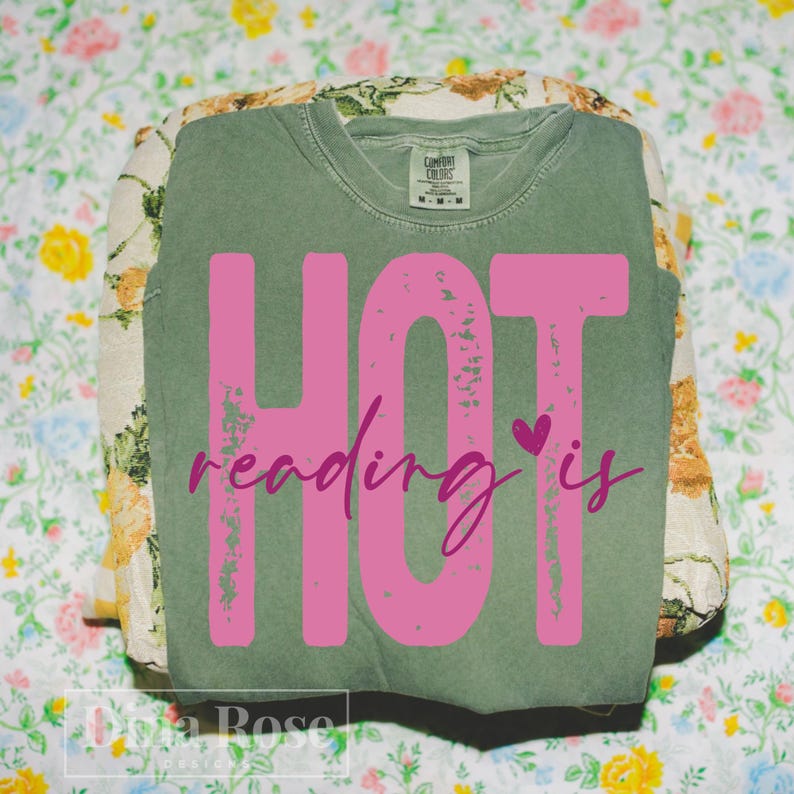 May include: A sage green Comfort Colors sweatshirt with the text "HOT reading is" in bright pink. The word "HOT" is in large block letters, and "reading is" is in a cursive font with a small heart. The shirt is folded on top of a floral patterned fabric.