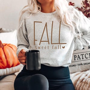 May include: A light gray sweatshirt with the word "FALL" in brown lettering, with "sweet fall" underneath. A black mug is held in the hand. A pumpkin and a pillow are in the background.