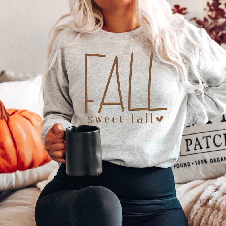 May include: A light gray sweatshirt with the word "FALL" in brown lettering, with "sweet fall" underneath. A black mug is held in the hand. A pumpkin and a pillow are in the background.