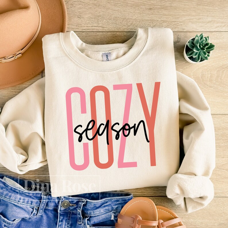 May include: A cream colored sweatshirt with the text "cozy season" in pink, red, and black lettering.