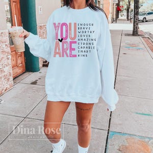May include: White sweatshirt with the text "YOU ARE" in pink and black letters. The text is surrounded by a list of positive words including "enough", "brave", "worthy", "loved", "amazing", "strong", "capable", "smart", and "kind".