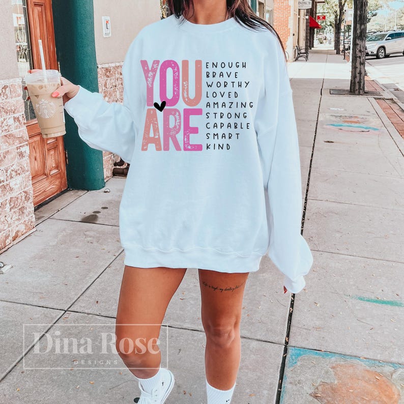 May include: White sweatshirt with the text "YOU ARE" in pink and black letters. The text is surrounded by a list of positive words including "enough", "brave", "worthy", "loved", "amazing", "strong", "capable", "smart", and "kind".