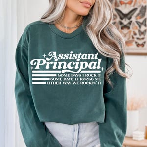 May include: Dark green sweatshirt with the text "Assistant Principal" in white script and block letters. Below the title are the words "SOME DAYS I ROCK IT, SOME DAYS IT ROCKS ME, EITHER WAY WE ROCKIN' IT" in white. A casual top for school staff.