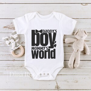 Peut inclure: Body de b&eacute;b&eacute; blanc avec l'inscription noire "Daddy's Boy, Mommy's World". Le body pr&eacute;sente un motif de n&oelig;ud papillon au-dessus du mot "boy" et deux c&oelig;urs. Un ours en peluche et un hochet en bois sont &agrave; c&ocirc;t&eacute; du body.