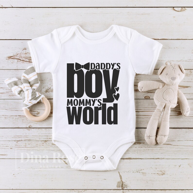 Peut inclure: Body de b&eacute;b&eacute; blanc avec l'inscription noire "Daddy's Boy, Mommy's World". Le body pr&eacute;sente un motif de n&oelig;ud papillon au-dessus du mot "boy" et deux c&oelig;urs. Un ours en peluche et un hochet en bois sont &agrave; c&ocirc;t&eacute; du body.