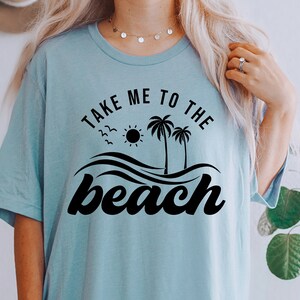 May include: A light blue t-shirt with the text "Take me to the beach" in black lettering. The design includes palm trees, a sun, and waves.
