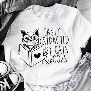 K&ouml;nnte beinhalten: Ein wei&szlig;es T-Shirt mit einer schwarz-wei&szlig;en Illustration einer Katze, die ein Buch liest. Der Text "Easily Distracted by Cats & Books" ist auf das T-Shirt gedruckt.